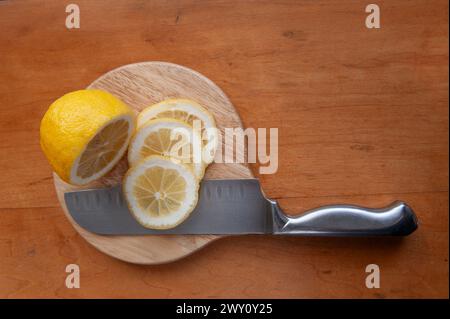 Drei frische Zitronenscheiben und eine halbe Zitrone auf einem runden hölzernen Schneidebrett mit einem Edelstahlmesser. Stockfoto