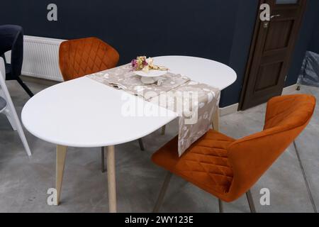 Weißer Holztisch kombiniert mit orangefarbenen Stühlen mit weichen Stoffpolstern. Es gibt eine weiße Lieferung mit einem mehrfarbigen Blumenstrauß auf dem t Stockfoto