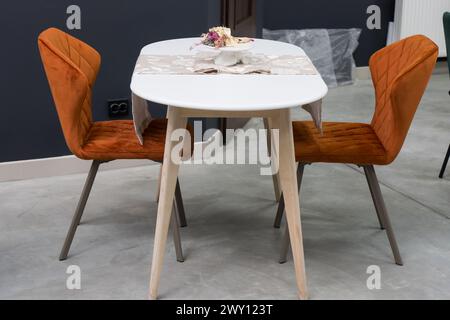 Weißer Holztisch kombiniert mit orangefarbenen Stühlen mit weichen Stoffpolstern. Es gibt eine weiße Lieferung mit einem mehrfarbigen Blumenstrauß auf dem t Stockfoto
