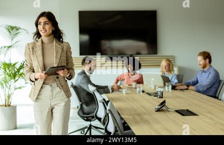 Entschlossene Führungskraft steht mit einem Tablet im Vordergrund und spricht ihr aufmerksames Team während eines Meetings an. Stockfoto