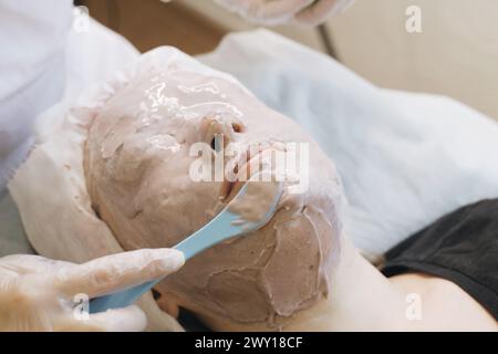 Eine Frau entspannt sich, während eine Spa-Therapeutin eine dicke Tonmaske auf ihr Gesicht aufträgt, die alles außer Augen und Mund für die Hautpflege bedeckt. Stockfoto