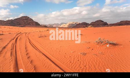 Felsige Massiven in der rot-orangen Sandwüste, Fahrzeugspuren, heller bewölkter Himmel im Hintergrund, typische Landschaft in Wadi Rum, Jordanien Stockfoto