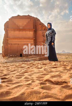 Frau in langen dunklen Kleidern posiert vor Mada'in Salih Lonely Castle oder Qasr al-Farid in Hegra, Saudia Arabien, sandige Wüstenlandschaft. Saudi Ar Stockfoto