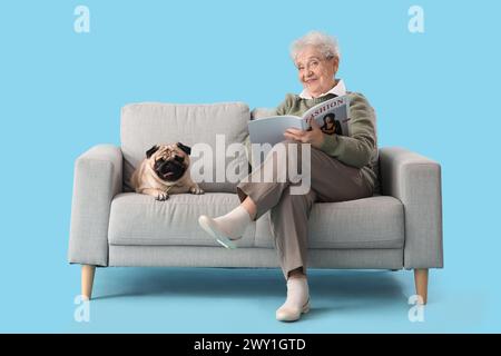 Ältere Frau mit Mops-Hund, der Zeitschrift auf Sofa vor blauem Hintergrund liest Stockfoto