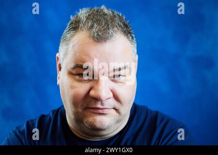 Unter seinen Augenbrauen blickt der 47-jährige Kaukasier mit leicht abgesenktem Kopf, das Gesicht ist dicht auf blauem Hintergrund. Stockfoto