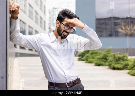 Müder und erschöpfter junger indischer Geschäftsmann, der draußen in der Nähe von Büros stand und seine Hand hielt, Schmerzen und Schwindel fühlte. Stockfoto
