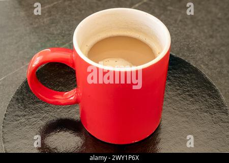 Rote Tasse Kaffee auf einem schwarzen reflektierenden Untersetzer und dunklem Tisch. Kaffee ist in der Tasse zu sehen. Stockfoto