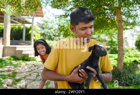 Ein entzückendes Bild eines indischen Vaters und seiner Tochter, die glückliche Erinnerungen schaffen, während sie mit einer charmanten jungen Ziege in ihrem lebhaften Hinterhof gar spielen Stockfoto