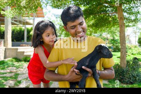 Wunderbares Bild eines indischen Vaters und seiner Tochter, die glückliche Erinnerungen beim Spielen mit einer charmanten jungen Ziege schaffen Stockfoto