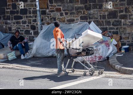 Kapstadt, Südafrika - 2. April 2024: Ein Obdachloser in einem orangefarbenen Hemd schiebt einen Einkaufswagen mit verschiedenen Gegenständen auf eine gepflasterte Fläche Stockfoto
