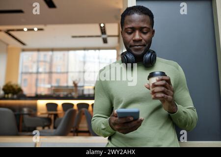 Porträt eines jungen afroamerikanischen Mannes mit einem Smartphone, das sich an der Wand in der Bürolounge beugt und Kaffee trinkt Stockfoto