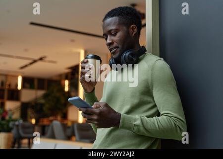 Minimales seitliches Porträt des jungen Schwarzen Mannes mit Smartphone, das sich an der Wand in der Bürolounge stützt und einen Kopierraum für Kaffee verwendet Stockfoto