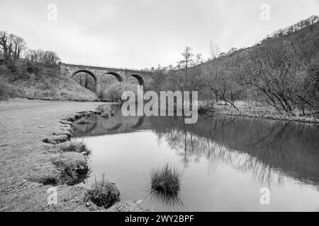 Ein Schwarzweiß-Bild der Monsal Head Bridge, die sich im Fluss Wye spiegelt, unter einem bewölkten Himmel. Stockfoto