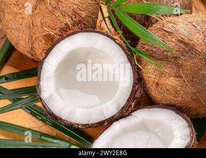 Frisch geöffnete Kokosnüsse zusammen mit ganzen Kokosnüssen und Kokosblättern auf einem Holztisch. Schöner exotischer Hintergrund für Ihre Projekte. Stockfoto
