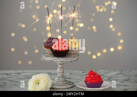 Verschiedene bunte Cupcakes mit Glitzern auf weißem Marmortisch vor verschwommenem Licht Stockfoto