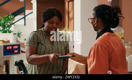 Weibliche Käuferin kauft ökologisch lokal angebaute Waren an der Kasse mit einer Karte am POS-System. Der Kunde wählt aus einer Vielzahl von Bio-Kulturen, Massenware oder Küchenartikeln in einem lokalen Geschäft. Stockfoto