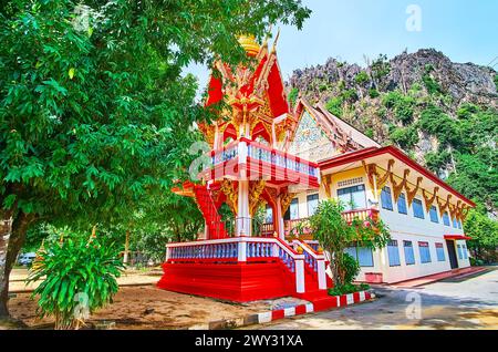 Beeindruckender hellroter Ho Rakang Glockenturm des Wat Suwan Kuha Höhlentempels hinter dem üppig grünen Baum, Phang Nga, Thailand Stockfoto