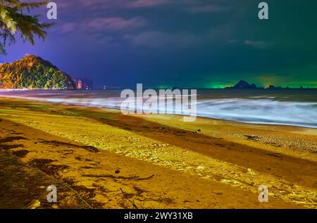 Romantischer nächtlicher Spaziergang am Sandstrand mit Blick auf die felsigen Inseln am Horizont und die hellen Lichter der Touristenboote, Ao Nang, Krabi, Thailand Stockfoto
