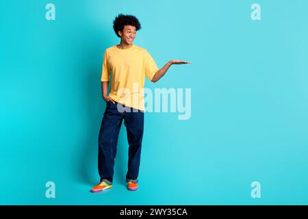 Foto in voller Größe von guter Stimmung Mann tragen übergroßes T-Shirt Denim Hosen Blick auf Objekt auf Arm leeren Raum isoliert auf blaublauem Hintergrund Stockfoto