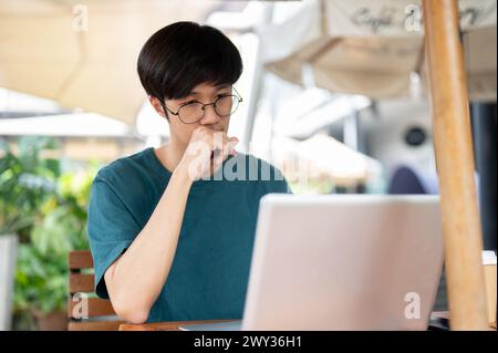 Ein nachdenklicher, seriöser junger asiatischer Mann mit Brille arbeitet ferngesteuert in einem Café in der Stadt und arbeitet an seinem Laptop, während er in einem Outdo sitzt Stockfoto