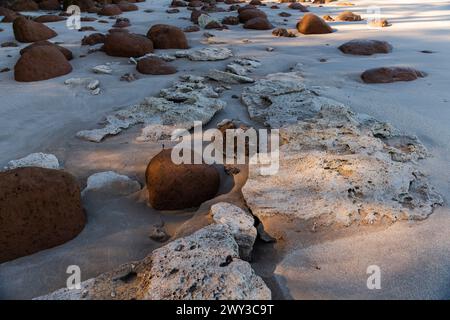 Ein großer Felsen liegt an einem Strand, umgeben von kleinen Felsen. Der Strand ist felsig und der Himmel ist klar Stockfoto