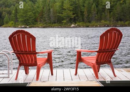 Zwei hellrote Adirondack-Plastikstühle auf hölzernem Schwimmsteg am ruhigen See mit Wald aus grünen Nadelbäumen und Laubbäumen in der späten Zeit Stockfoto