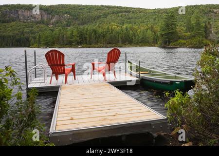 Zwei hellrote Adirondack-Plastikstühle auf einem schwimmenden Holzdock und ein grünes Ruderboot auf einem ruhigen See mit Wald aus grünen Nadelbäumen und Laubbäumen Stockfoto