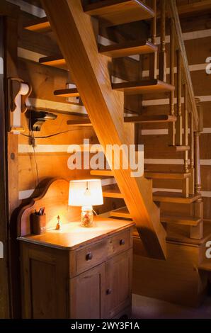 Kleiner antiker Holzschrank mit beleuchteter Lampe unter einer Holztreppe mit flachen Stufen, die von einem einzigen Balken gestützt werden und zum Boden im Obergeschoss führen Stockfoto