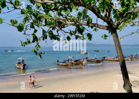 Ein westliches Touristenpaar, das am Ao Nang Beach, Krabi, Südthailand vorbeiläuft, vorbei an traditionellen thailändischen Booten, die am Ufer ankern Stockfoto
