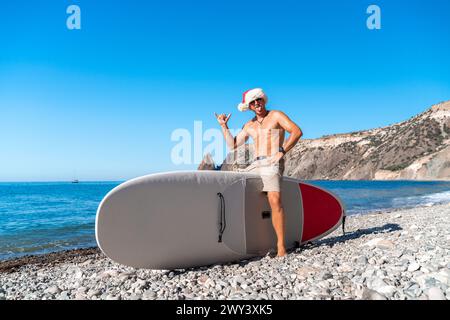 Ein Mann mit einem Weihnachtsmann-Hut sitzt auf einem umgedrehten SUP-Board am Meer. Er zeigt seine Zunge und hält seine rechte Hand hoch. Vor dem Hintergrund Stockfoto