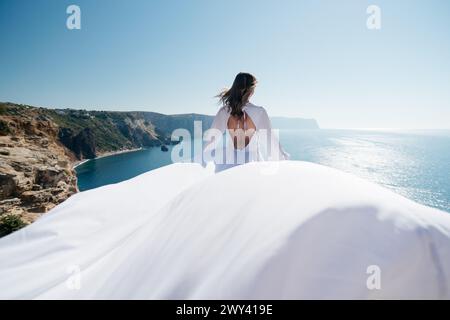 Eine Frau in einem weißen Kleid steht auf einer Klippe mit Blick auf den Ozean. Die Szene ist ruhig und friedlich, das Kleid der Frau fließt im Wind Stockfoto