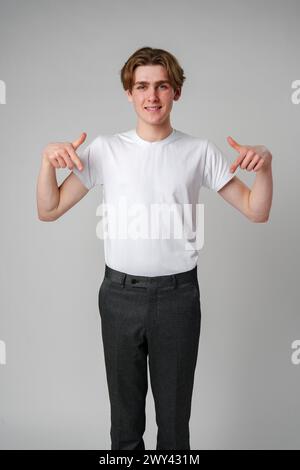 Junger Mann in weißem T-Shirt, das nach unten zeigt, mit beiden Händen vor glattem Hintergrund Stockfoto
