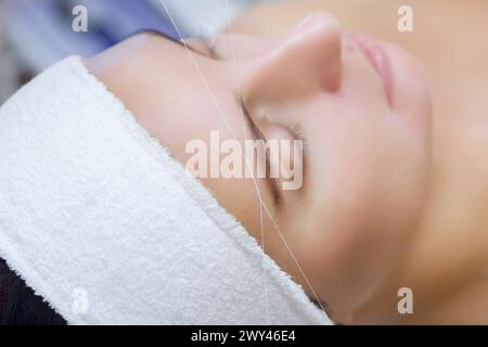 Die Make-up-Künstlerin zupft ihre Augenbrauen mit einer Fadennaht. Schönheitsbehandlungen für Gesichtspflege im Schönheitssalon. Stockfoto