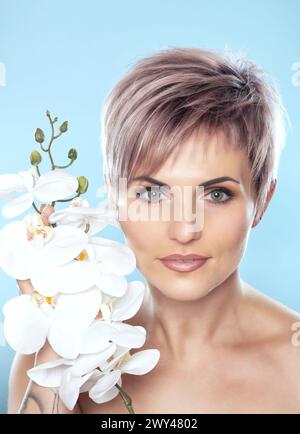 Porträt einer schönen blonden Frau mit weißer Orchidee in der Hand auf blauem Hintergrund. Professionelle Hautpflege und Make-up. Stockfoto