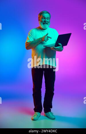 Voller Mann, der mit Laptop steht, lächelt und auf dem Bildschirm zeigt, auf blau-violettem Hintergrund im Neonlicht Stockfoto