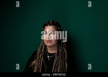 Eine Frau mit Dreadlocks steht zuversichtlich vor einer leuchtend grünen Wand, ihre Haare kaskadieren ihren Rücken hinunter. Sie sieht direkt in die Kamera, Exsudi Stockfoto