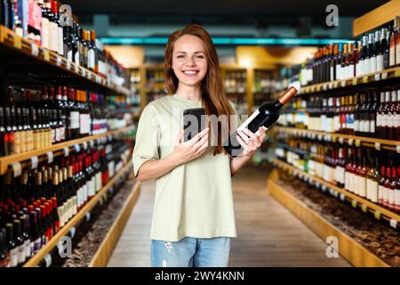 Glückliche junge Einzelhandelskunde wählt Wein in einem Weingeschäft. Frau steht im Weinladen und hält Weinflasche und Handy in der Hand. Stockfoto