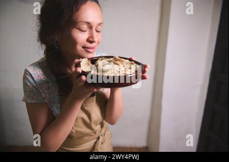 Authentisches Porträt einer schönen jungen brünetten Frau in beigefarbener Küchenschürze, lächelnd und genießt das Aroma frisch gekochter Knödel mit püriertem Topf Stockfoto