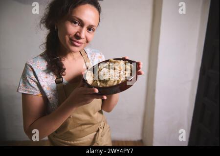 Attraktive junge Hausfrau in beigefarbener Kochschürze, lächelnd in die Kamera blickend, mit einem Tongericht mit frisch gekochtem, hausgemachtem ukrainischen Knödel Stockfoto