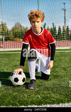 Ein kleiner Junge in einer Fußballuniform kniet anmutig auf dem Gras und hält einen Fußball vor sich. Stockfoto