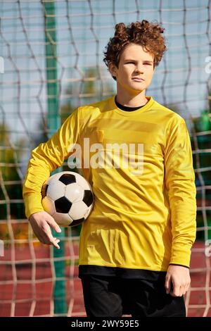 Ein junger Mann steht selbstbewusst vor einem Fußballnetz und hält einen Fußball. Er blickt nach vorne und ist bereit, sich der Herausforderung zu stellen, ein Tor zu erzielen. Stockfoto