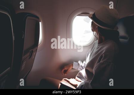 Frau schaut aus dem Fenster eines fliegenden Flugzeugs. Junge kaukasische glückliche Passagiere reisen mit dem Flugzeug, beobachten den Himmel von oben und halten Passpo Stockfoto