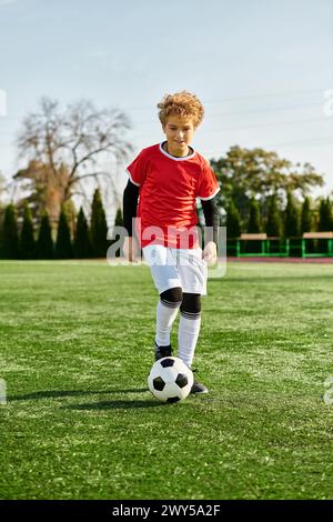 Ein kleiner Junge zeigt seine Fußballfähigkeiten, indem er selbstbewusst einen Fußball auf einem grasbewachsenen Feld kickt. Sein Fokus und seine Entschlossenheit scheinen durch, während er praktiziert Stockfoto