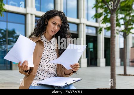 Besorgte junge Frau, die Dokumente mit einem verwirrten Ausdruck vor einem modernen Bürogebäude ansieht. Stockfoto