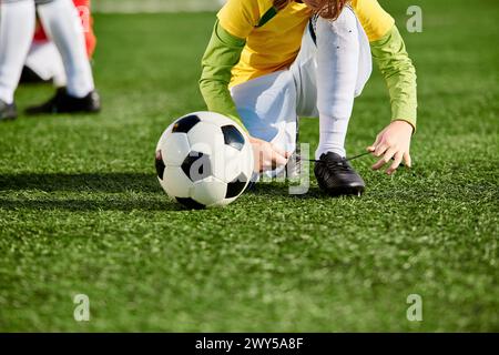 Ein junges Mädchen mit Zöpfen kniet auf einem Feld nieder und greift nach einem Fußball mit bunten Mustern. Stockfoto