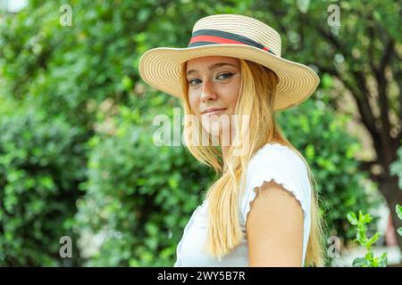 Eine blonde Frau mit Strohhut steht in einem Park. Sie hat ein Lächeln im Gesicht und blickt direkt in die Kamera Stockfoto