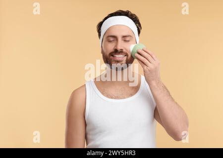 Mann mit Stirnband, der sein Gesicht mit einem Schwamm auf beigem Hintergrund wäscht Stockfoto