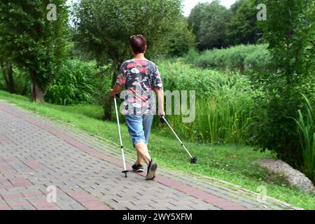 Das Bild zeigt eine ältere Dame, die mit Stöcken in der Hand im Park beim Sport geht. Stockfoto