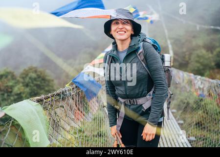 Junge lächelnde Backpackerin überquert den Canyon über die Hängebrücke mit bunten tibetischen Gebetsfahnen. Mera Gipfel Kletterroute Wanderung Lukla Saga Stockfoto