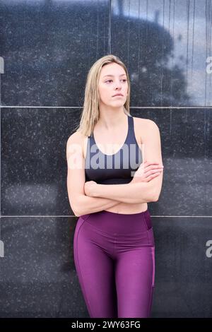 Schlanke blonde Frau in Sportbekleidung an einer Wand mit überkreuzten Armen. Vertikales Foto Stockfoto